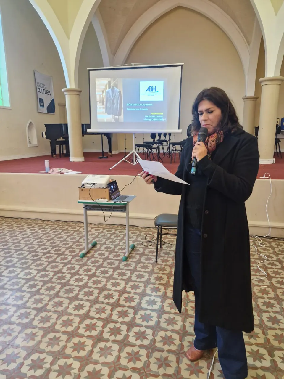 Palestrante ao microfone diante da apresentação institucional da ABIH-RJ projetada no telão